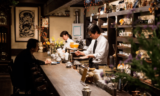 cafeteria kissaten típica, ambiente intimista, prateleiras de madeira ao fundo da área dos baristas repleta de xícaras japonesas, um homem e uma mulher preparando café e outro sentado em banqueta aguardando o café