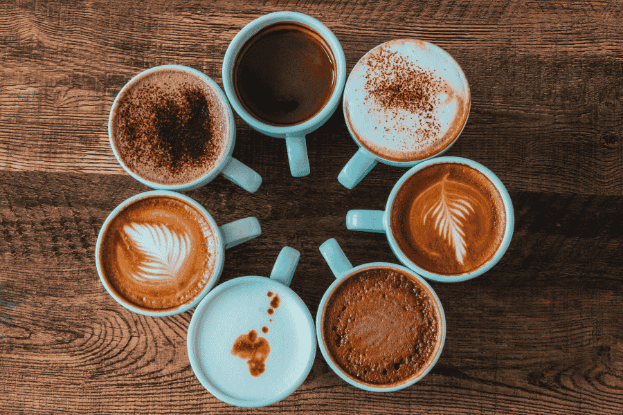 sobre uma mesa de madeira, 7 xícaras de café com diferentes tipos de café formando um círculo
