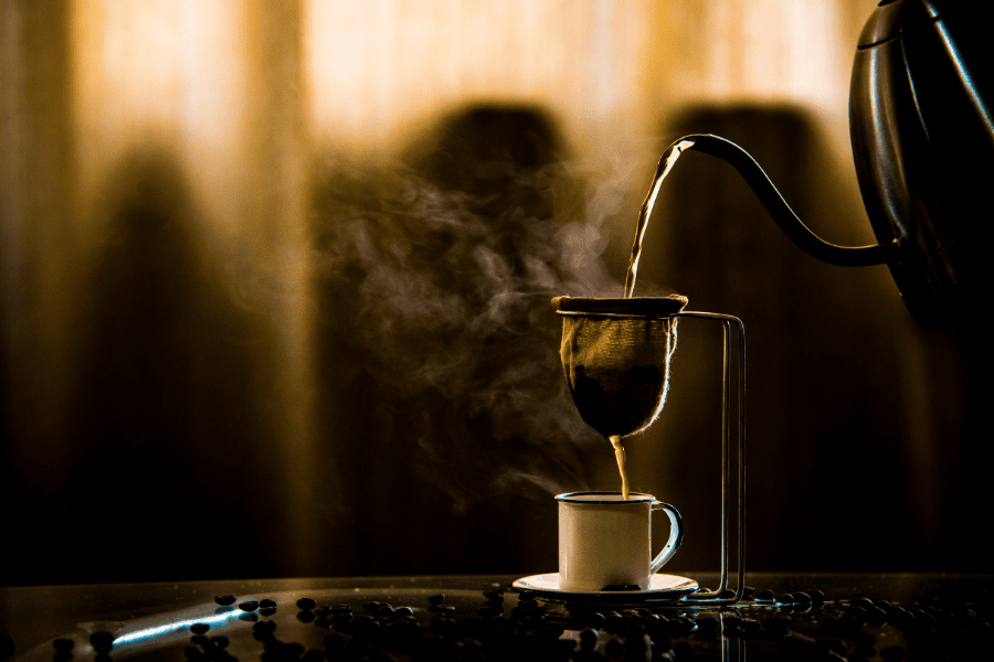 sobre uma mesa de vidro, uma xícara de louça de café abaixo de um passador de pano. Uma chaleira serve água e passa o café na xícara