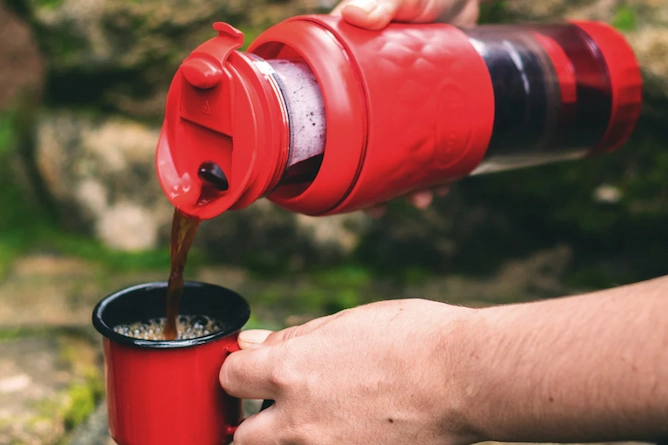 em um ambiente externo como uma trilha, uma pessoa está servindo café com uma Pressca, método de preparo brasileiro. Ela é de plástico com detalhes em vermelho, uma cafeteira portátil semelhante a prensa francesa. Serve em uma xícara de metal vermelha