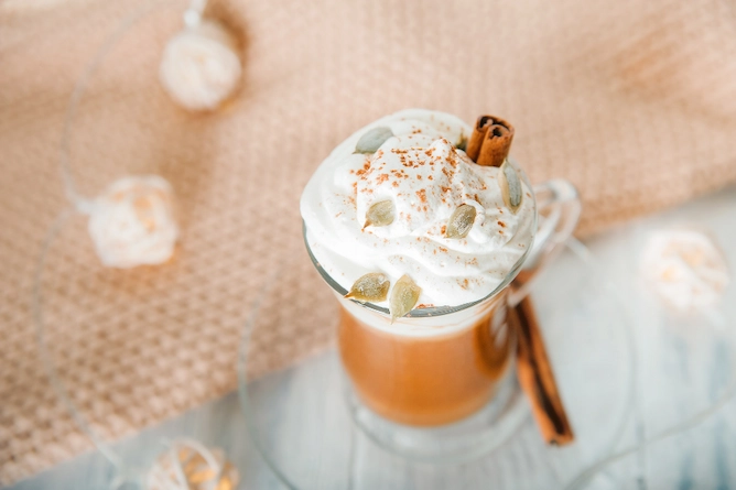 sobre uma mesa, uma toalha de tecido trançado e ao lado, uma xícara de vidro com a receita servida, finalizada com chantilly, canela em pau e em pó e outras especiarias