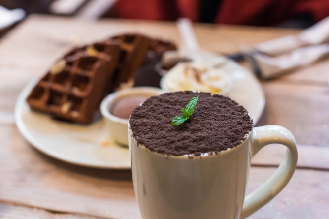 sobre uma mesa de madeira, ao fundo um prato com waffles e sorvete e no plano da frente, uma xícara cheia de café finalizado com raspas de café e folhas de hortelã