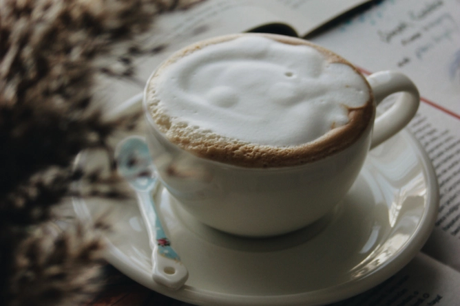 sobre um livro, um pires com uma colher pequena e uma xícara de porcelana cheia de café com leite cremoso