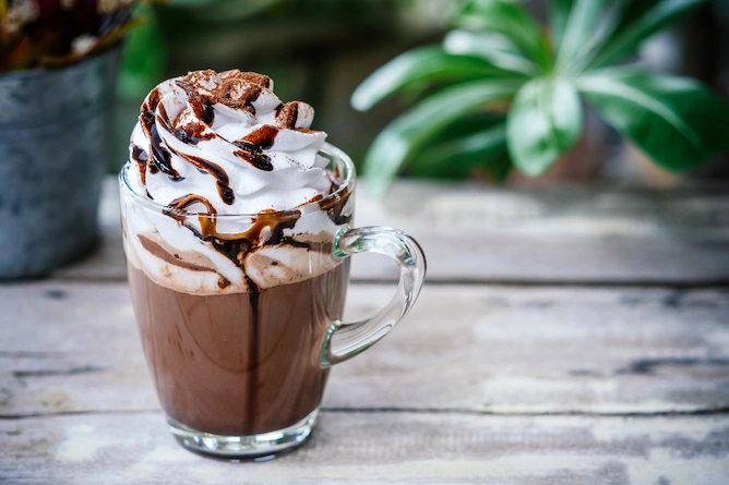 sobre uma mesa de madeira, com plantas no fundo, uma xícara de vidro com chocolate quente, finalizado com chantilly e uma espécie de calda de chocolate