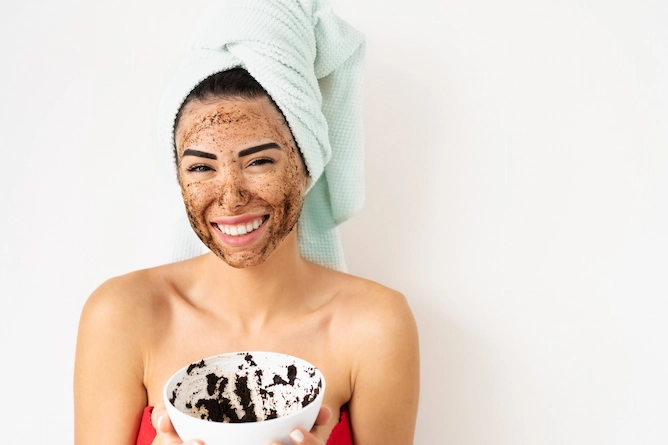 mulher sorridente com toalha enrolada no cabelo, com máscara de café no rosto segurando uma vasilha que contém a mistura da máscara