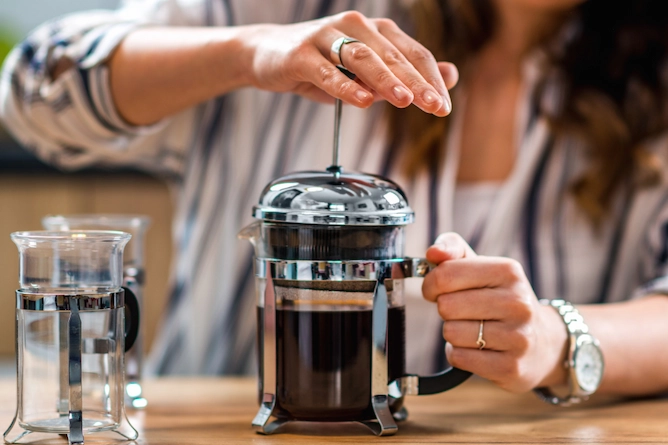 em uma mesa de madeira, a mulher abaixa o êmbolo da prensa francesa (cafeteira de vidro com detalhes em inox)