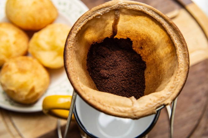 em uma mesa, um prato com pães de queijo e em primeiro plano, um coador de pano coando café moído