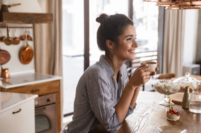 uma mulher sentada numa mesa da cozinha, sorridente, segurando uma xícara de café nas mãos