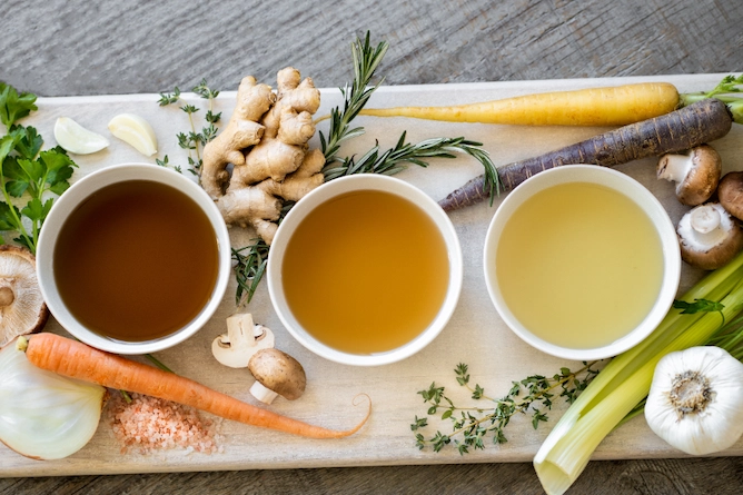 tábua de madeira com legumes, cenouras, cogumelos, alho, gengibre, alecrim e três caldos de legumes