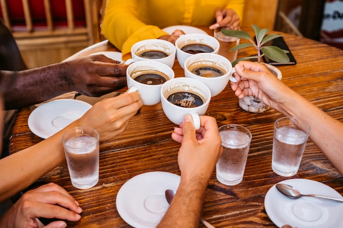 sobre uma mesa de madeira redonda, cinco amigos brindam com xícaras de café. Na mesa, ainda têm copos d`água com gás para acompanhar, os pires das xícaras e colheres de chá