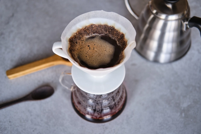 método de preparo V60 ou Hario passando café em filtro de papel, sobre uma mesa, com uma chaleira de inox atrás e colheres ao lado