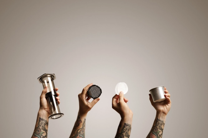quatro mãos segurando as quatro partes do equipamento: estrutura plástica, êmbolo, filtro de papel e recipiente para o café