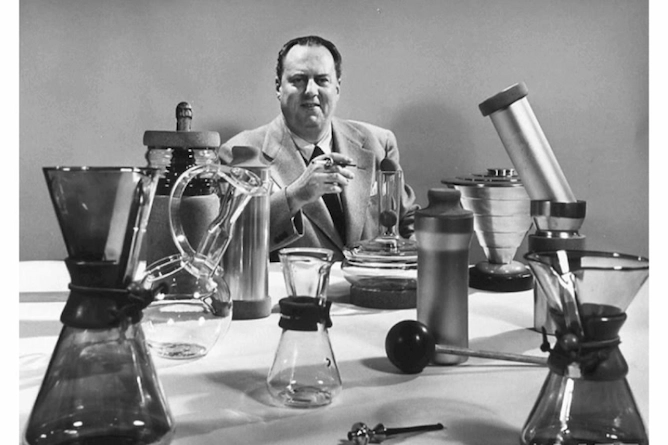 criador da chemex sentado em uma mesa com muitos equipamentos teste da cafeteira, muitos tipos de vidraçarias, parecendo um laboratório