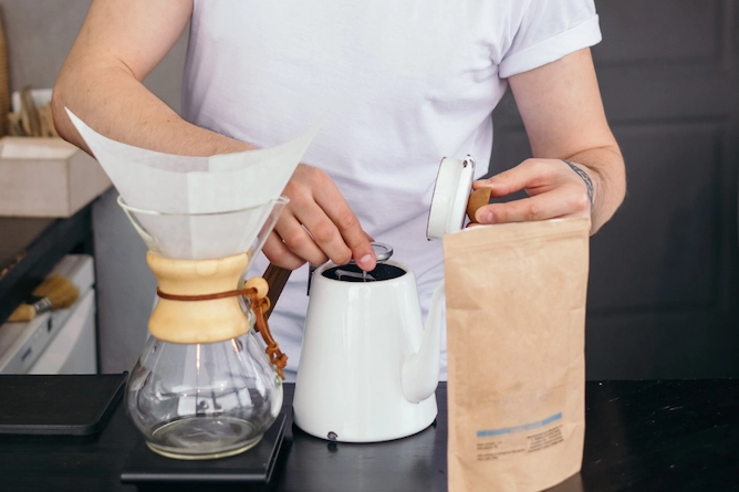 sobre uma bancada de cozinha, uma chemex sobre uma balança, ao lado uma chaleira e um pacote de café