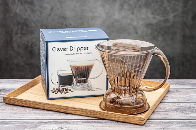 sob uma mesa de madeira, em cima de uma bandeja retangular, uma caixa da clever (o produto) e ao lado a estrutura de acrílico da cafeteira