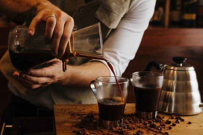 sobre uma mesa de madeira, uma chaleira bico de ganso e duas xícaras transparentes de café, sendo servido em uma delas um café feito na chemex