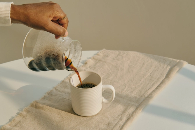 sobre uma mesa de madeira, um pano de algodão e uma caneca de porcelana onde se está sendo servido café