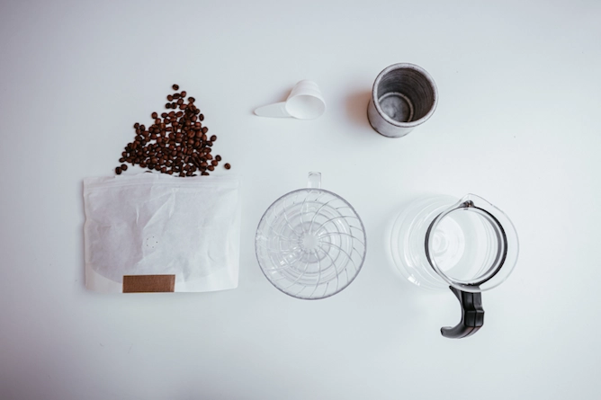em cima de uma mesa, um pacote de café em grãos com alguns grãos caindo para fora, o suporte V60, uma colher medidora, um copo e um vidro (o recipiente do V60)