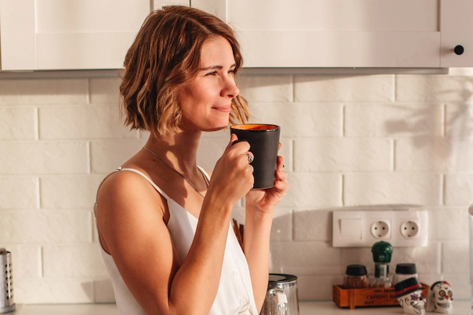 mulher com cabelos curtos encostada em um balcão de cozinha, segura uma caneca de café com semblante feliz e tranquilo