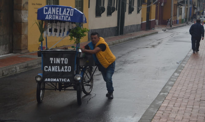 Vendedor empurra triciclo azul escrito “Tinto” e “Canelazo” em rua molhada de Bogotá, prédios coloniais ao fundo.